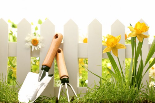 A close-up image of a small garden scene featuring a white wooden picket fence with yellow daffodils and white daisies growing in front of it, set on a lush green lawn. In the foreground, there are two gardening tools—a trowel and a pair of pruning shears—resting on the grass, indicating active gardening or maintenance work. The background shows a soft, out-of-focus greenery, suggesting a well-maintained outdoor space that could be part of a residential garden in Bayswater, London. The natural daylight and mild weather conditions highlight the vibrant colours of the flowers and the fresh, healthy appearance of the lawn, aligning with gardening services offered by Landscapers Bayswater for garden enhancement and patio repair solutions in the local area.