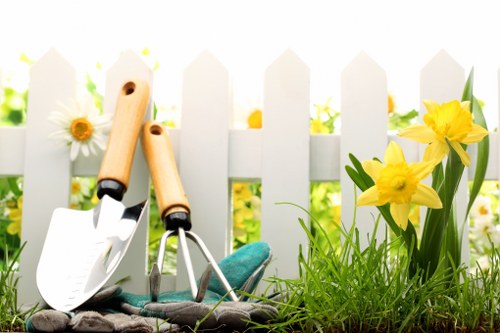 The image depicts a close-up view of a small garden area with lush green grass and blooming yellow daffodils situated near a white wooden picket fence. In the foreground, there are gardening tools including a trowel with a wooden handle, a small hand fork, and a pair of gardening gloves, all resting on the ground next to the plants. The background features the white fence with sunlight filtering through, creating a bright and inviting outdoor environment typical of well-maintained residential gardens. This scene highlights features common in gardening and outdoor maintenance, such as cultivating flower beds and maintaining lawn areas, which are services offered by companies like Landscapers Bayswater. The overall setting suggests a neat, organized, and thriving garden space suitable for various landscaping and gardening projects in Bayswater or surrounding areas, emphasizing the natural tones and textures of the soil, plants, and garden tools under soft natural light.