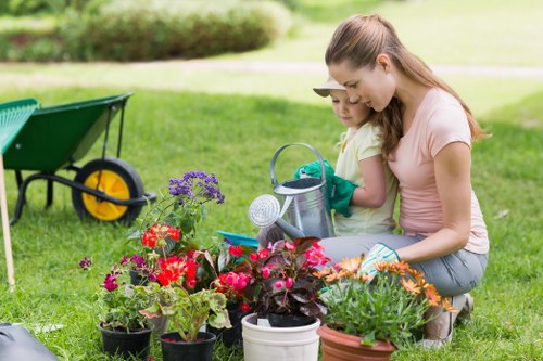 A woman and a young girl are gardening together in an outdoor garden space in Bayswater, surrounded by various potted flowering plants, including red, purple, and orange blooms, placed on lush, green grass. The woman, with light brown hair tied back, is wearing a pink T-shirt and jeans, while the girl has blonde hair and is dressed in a yellow T-shirt. They are engaged in planting or watering activities, with small gardening tools and a watering can nearby. In the background, there are trees, a pathway, and a small, green garden wheelbarrow with a yellow wheel, indicating a well-maintained outdoor space suitable for gardening and outdoor maintenance services by Landscapers Bayswater. The scene is lit by natural daylight, highlighting the vibrant colours of the plants and the natural textures of the grass and garden elements, conveying a peaceful and productive gardening environment in a typical residential outdoor area in Bayswater.